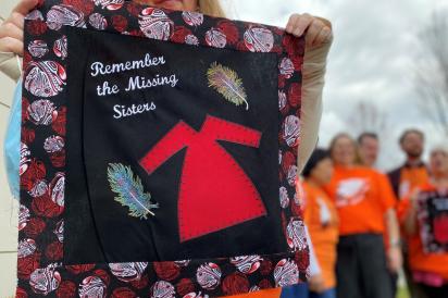 Member holding up quilt square with words "Remember the missing sisters". HEU members with orange t-shirts in the background